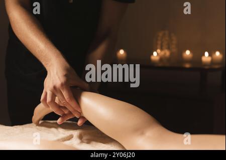 Primo piano di mani maschili che fanno un massaggio polpaccio di gambe femminili in una stanza buia con candele sullo sfondo. Trattamenti cosmetici e spa Foto Stock