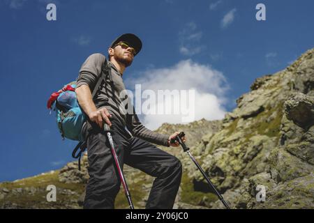 Porter di un elegante viaggiatore hipster con una barba e uno zaino in occhiali da sole e un berretto con pali da trekking in piedi su una roccia sullo sfondo Foto Stock