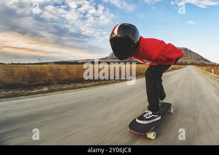 Un giovane con casco integrale e una tuta in pelle in uno speciale rack, cavalca un longboard sul tramonto afsaltu sulle montagne sullo sfondo e sul bellissimo sk Foto Stock