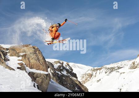 Uno sciatore professionista fa un salto da un'alta scogliera contro il cielo blu lasciando una scia di neve in montagna. Foto dalle piste o Foto Stock