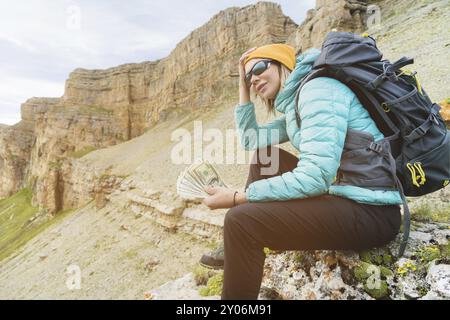 Una ragazza viaggiatrice che indossa un cappello e occhiali da sole tiene un biglietto da cento dollari nelle mani di un fan sullo sfondo di scogliere sulla natura. Tenga il suo lui Foto Stock