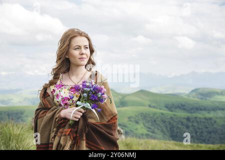 Una bella ragazza bionda avvolta in un plaid si staglia con un bouquet di fiori selvatici e guarda lo sguardo sullo sfondo di un bellissimo paesaggio naturale Foto Stock