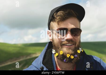 Ritratto ravvicinato di un uomo allegro barbuto con occhiali da sole e un cappello grigio con fiori selvatici in barba. Brutalità morbida e buona mascolinità Foto Stock