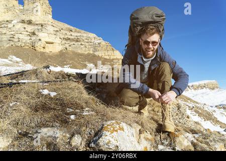 Ritratto di un viaggiatore hipster che ride felice con la barba in occhiali da sole è seduto e legando lacci di scarpe alla natura. Un uomo che cammina in montagna Foto Stock