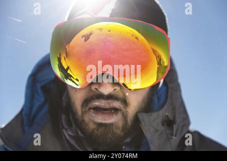 Un ritratto di un uomo barbuto che indossa occhiali da sci mentre si dirige verso la cima della montagna. Il concetto di resistenza nel cammino sullo sfondo della montagna Foto Stock