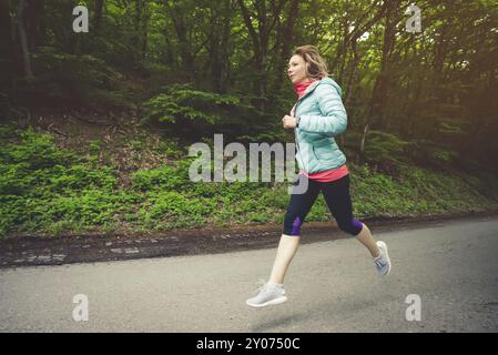 Giovane donna bionda fitness in cuffia che corre al mattino percorso forestale caucasico in luce solare Foto Stock