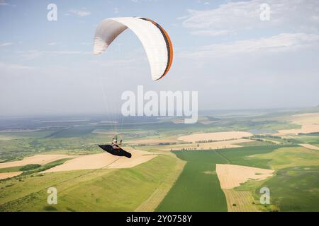 Il parapendio professionista in tuta di cocco vola in alto sopra il terreno contro il cielo e i campi Foto Stock