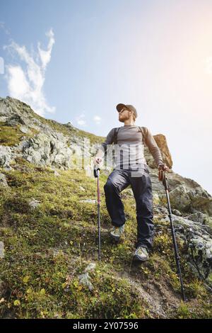 Un uomo barbuto con occhiali da sole e un berretto con uno zaino si staglia sulla cima di una roccia e guarda verso una valle rocciosa in alto tra le montagne. Il concetto di tour Foto Stock