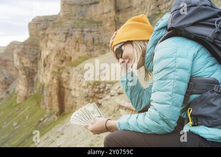 Una ragazza viaggiatrice che indossa un cappello e occhiali da sole tiene un biglietto da cento dollari nelle mani di un fan sullo sfondo di scogliere sulla natura. Tenga il suo lui Foto Stock