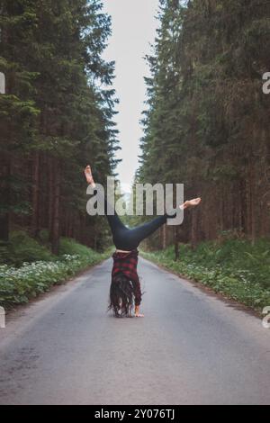 Donna acrobatica con leggings neri e camicia a quadri esegue un cartoncino su un sentiero nella foresta nel mezzo di una foresta profonda. Formazione di coordinamento e agilità Foto Stock