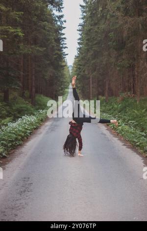 Donna acrobatica con leggings neri e camicia a quadri esegue un cartoncino su un sentiero nella foresta nel mezzo di una foresta profonda. Formazione di coordinamento e agilità Foto Stock