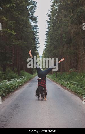 Donna acrobatica con leggings neri e camicia a quadri esegue un cartoncino su un sentiero nella foresta nel mezzo di una foresta profonda. Formazione di coordinamento e agilità Foto Stock