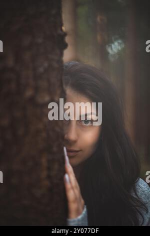 Una donna enigmatica con gli accattivanti occhi blu guarda da dietro un albero in una foresta mistica. L'immagine cattura l'essenza del mistero, della natura e del seren Foto Stock