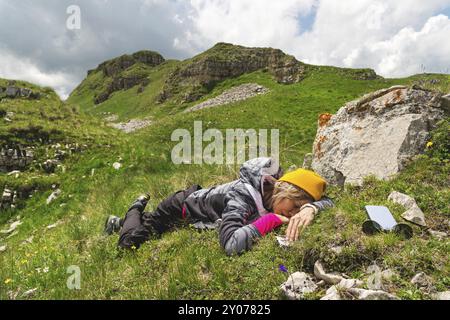 Ragazza all'aperto che dorme sull'erba verde contro una pietra in natura. Pericolo e fatica in natura Foto Stock