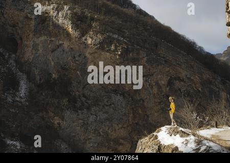 Ritratto a figura intera Una fotografa con occhiali da sole e un cappello di pelliccia grande e un maglione giallo in maglia si staglia sullo sfondo di alte rocce in t Foto Stock