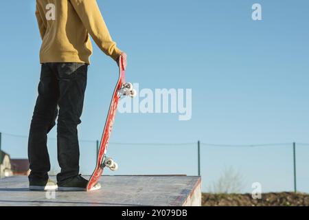 Primo piano Un adolescente in piedi con una felpa gialla tiene uno skateboard a mano sullo sfondo della periferia della baraccopoli e del cielo blu della città Foto Stock