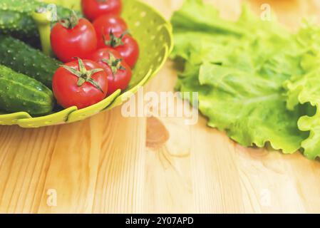 Verdure fresche, cetrioli e pomodori in un recipiente verde su un tavolo di legno accanto alle foglie di lattuga verde. Dieta vegetariana. Dieta sana e stile di vita Foto Stock