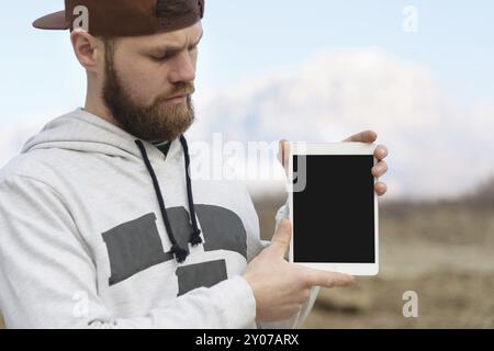 Primo piano Ritratto di un hipster in un berretto marrone all'aperto tiene in mano un tablet pc bianco. Un uomo barbuto guarda la tavoletta. sfocatura dello sfondo Foto Stock
