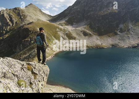 Un turista hipster barbuto in occhiali da sole con uno zaino è in piedi sul bordo di una scogliera in cima alle montagne vicino al lago di montagna Foto Stock