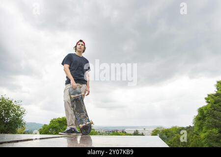 Un giovane pattinatore a pelo lungo con una maglietta e un berretto a tutta lunghezza è in piedi con uno skateboard sullo sfondo del paesaggio urbano Foto Stock