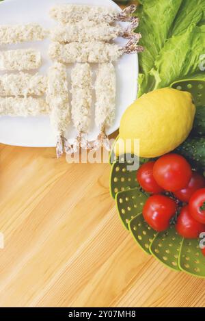 Gamberi crudi in pastella pronti per la tostatura accanto a un cestino di cetrioli e cetrioli di limone e pomodori sullo sfondo di foglie di lattuga su un legno Foto Stock