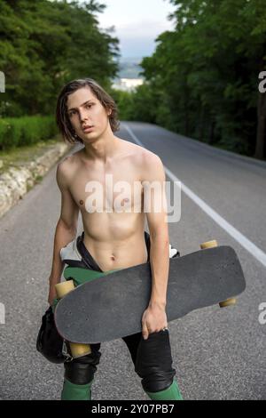 Un giovane uomo con un abito in pelle e capelli lunghi sta sull'asfalto con un casco tra le mani e il suo longboard sullo sfondo del verde e. Foto Stock