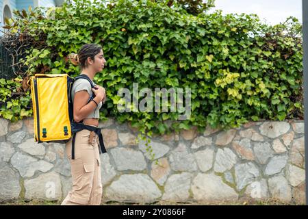 Vista laterale da copyspace di una ragazza ispanica che consegna cibo con uno zaino giallo mentre cammina attraverso un quartiere residenziale per consegnare gli ordini. L'ima Foto Stock