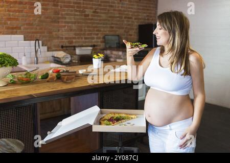 Giovane donna incinta di mangiare la pizza alla cucina. Nutrizione in gravidanza Foto Stock