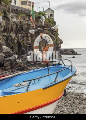 Una colorata barca da pesca con salvagente su una costa rocciosa vicino al mare, Funchal, madeira, portogallo Foto Stock