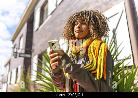 Giovane donna latina che usa il telefono in piedi in una strada residenziale indossando abiti caldi e sciarpa in una giornata di sole Foto Stock