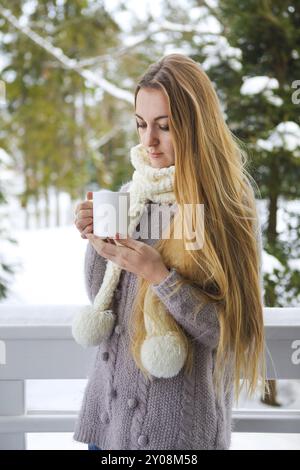 Giovane e bella donna con capelli biondi all'aperto e tazza di caffè caldo con maglione caldo lavorato a maglia. Concetto di comfort invernale Foto Stock