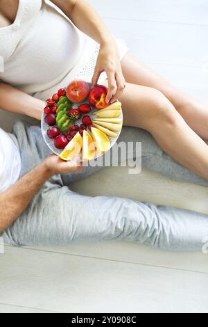 Giovane uomo e donna incinta seduta con un piatto di frutta. La gravidanza e il concetto di nutrizione Foto Stock