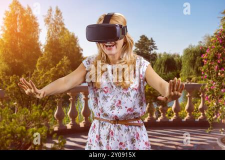 Donna matura felice e stupita con un visore per realtà virtuale in piedi all'aperto che si allunga le braccia. Parco cittadino sullo sfondo. Foto Stock
