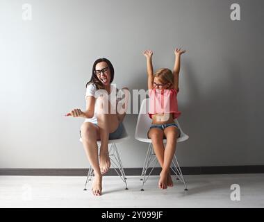 Felice sua madre e la sua piccola figlia seduti sulle sedie dal muro grigio. Ritratto di Madre e figlia con occhiali Foto Stock