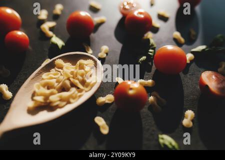 Pomodori ciliegini rossi, basilico verde, vermicelli a farfalla, cucchiaio di legno su sfondo grigio. Foto Stock