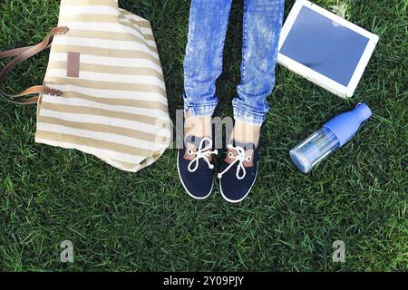 Piedi di una giovane ragazza su uno sfondo di un prato verde con bottiglia di tablet PC, di acqua e di borsa. Viaggi e concetto di holliday Foto Stock