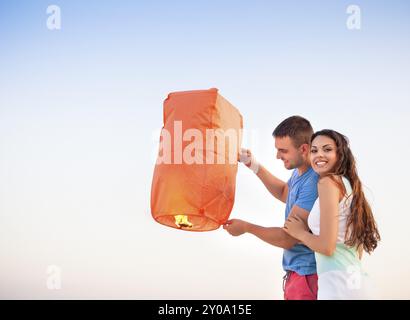 Coppia giovane iniziare un rosso cielo cinese lanterna nel tramonto vicino al mare Foto Stock