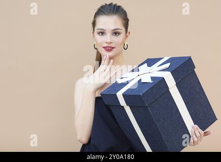 Sorpreso ragazza carina in blu navy vestiti azienda big box regalo isolato sul beige Foto Stock