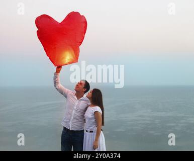 Coppia giovane iniziare un rosso cielo cinese lanterna nel tramonto vicino al mare Foto Stock