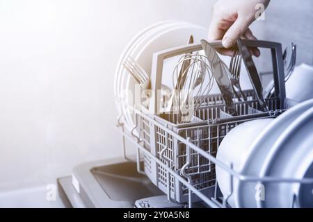 Primo piano della mano femmina che tiene il cestello completo delle posate con coltello pulito, forchetta, frusta. Caricamento, svuotamento o scaricamento dalla lavastoviglie automatica aperta m Foto Stock