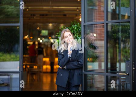 Una donna premurosa interessata a una tuta da lavoro che tiene il telefono in attesa all'entrata del caffè guardando da parte Foto Stock