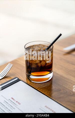 Cocktail di bibita a base di whiskey e cola al tavolo da pranzo del ristorante con menu per la cena e argenteria Foto Stock