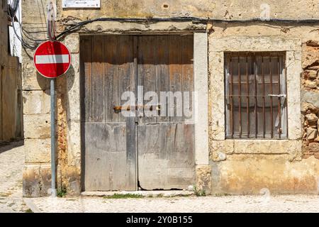 Vecchia porta in legno con serratura arrugginita e finestra barrata sono collocate in un muro di pietra, con un cartello di ingresso in primo piano Foto Stock