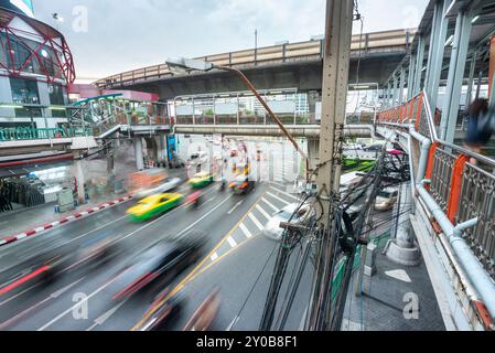 Movimento offuscato del traffico cittadino, che corre sotto un passaggio pedonale, mentre uno Sky-train sopraelevato e pieno di passeggeri attraversa rapidamente la congestione Foto Stock