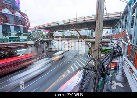 Movimento offuscato del traffico cittadino, che corre sotto un passaggio pedonale, mentre uno Sky-train sopraelevato e pieno di passeggeri attraversa rapidamente la congestione Foto Stock