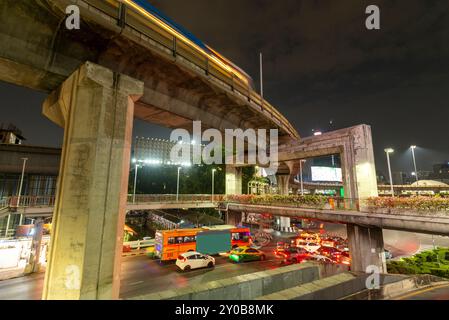 Il traffico in attesa di un incrocio affollato e molti veicoli corrono sotto enormi colonne di cemento, architettura che supporta l'estesa rete ferroviaria urbana di Bangkok. Foto Stock