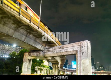 Di notte si avvicina un treno passeggeri sospeso, che corre su enormi pilastri di cemento, sostenendo le massicce ferrovie curve, che circondano una trafficata città cen Foto Stock