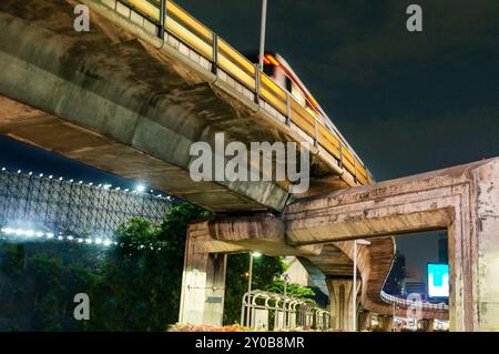 Di notte si avvicina un treno passeggeri sospeso, che corre su enormi pilastri di cemento, sostenendo le massicce ferrovie curve, che circondano una trafficata città cen Foto Stock