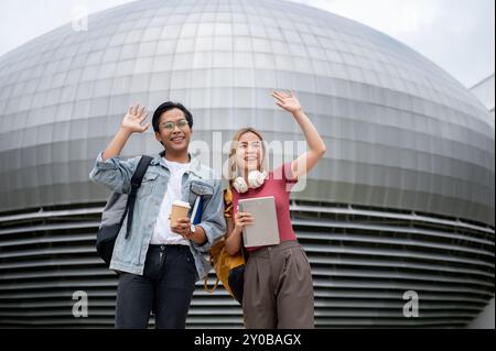 Due allegri giovani studenti universitari asiatici, un maschio e una femmina, in abiti casual e zaini, alzano le mani e sorridono mentre ti alzano Foto Stock