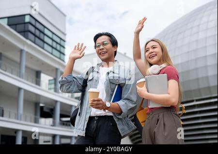 Due allegri giovani studenti universitari asiatici, un maschio e una femmina, in abiti casual e zaini, alzano le mani e sorridono mentre ti alzano Foto Stock